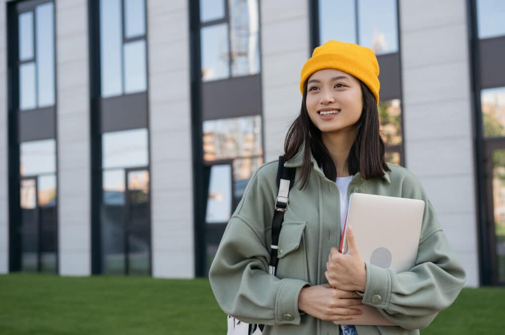 Smiling asian woman holding laptop computer looking away on the street. Happy student walking to university