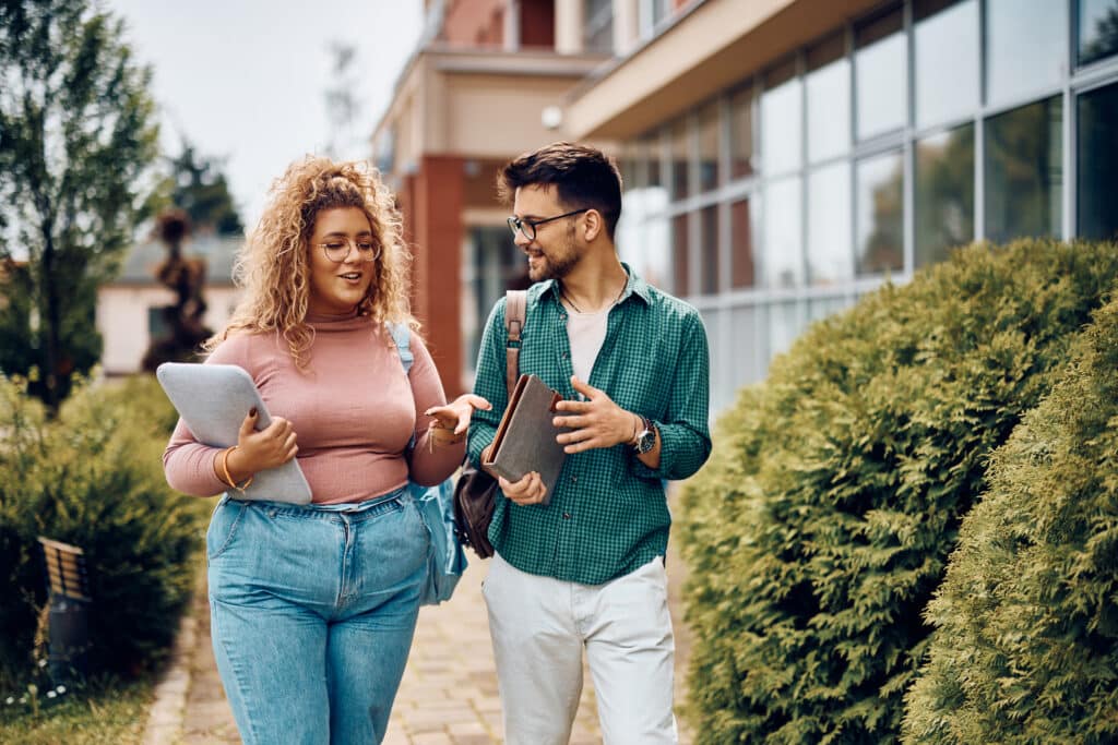 Happy student and her male friend communicating while walking through university campus.