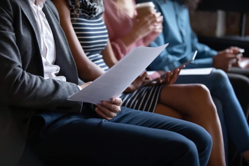 Close up unrecognizable young multiracial people sitting in line on sofa with paper documents or modern technology gadgets, waiting for job interview.