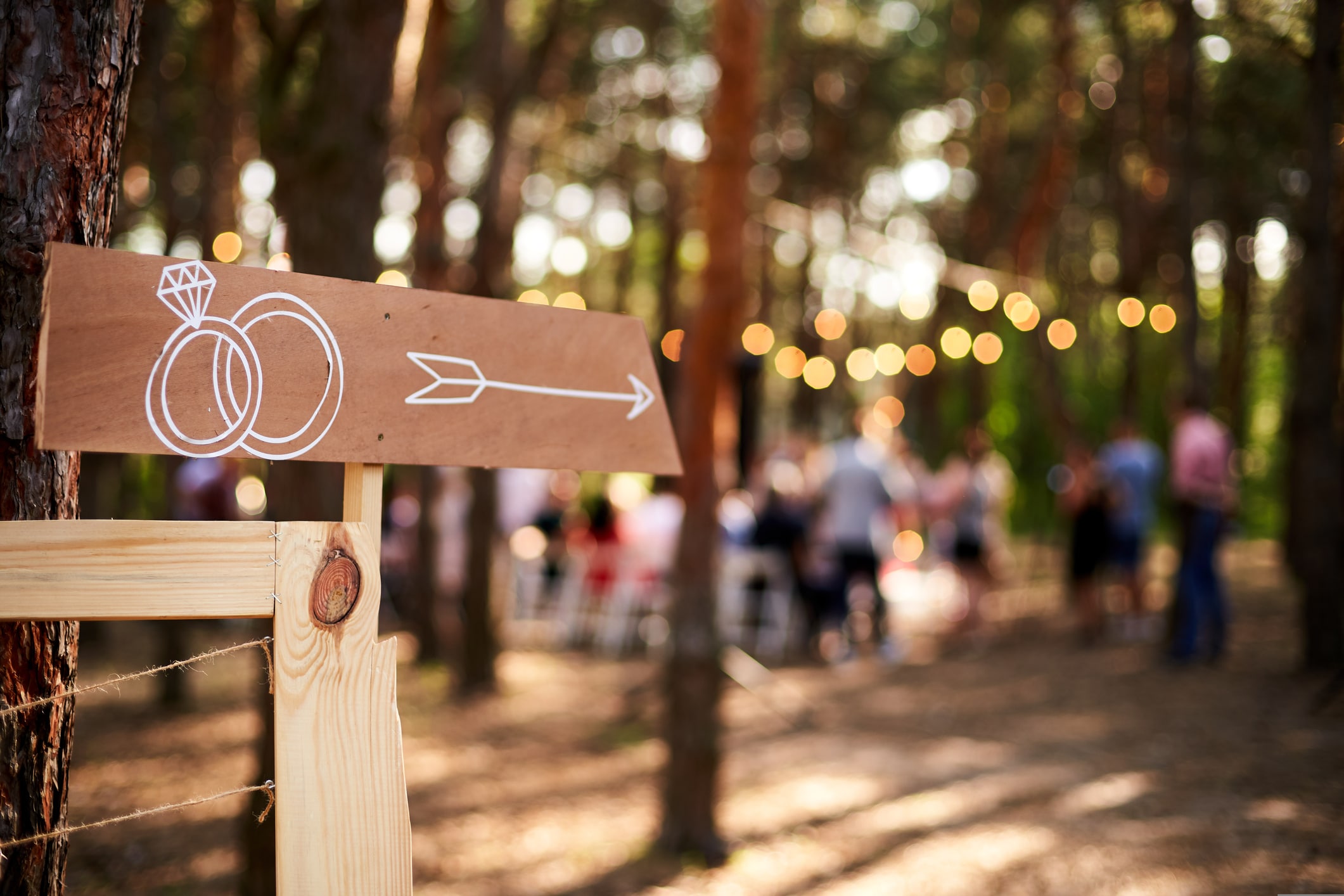 wedding ideas on a budget. Wooden arrow sign with wedding rings on ceremony venue. Wedding party banner post . Direction info banner for guests in pine forest outdoors. Rustic, country style decor.