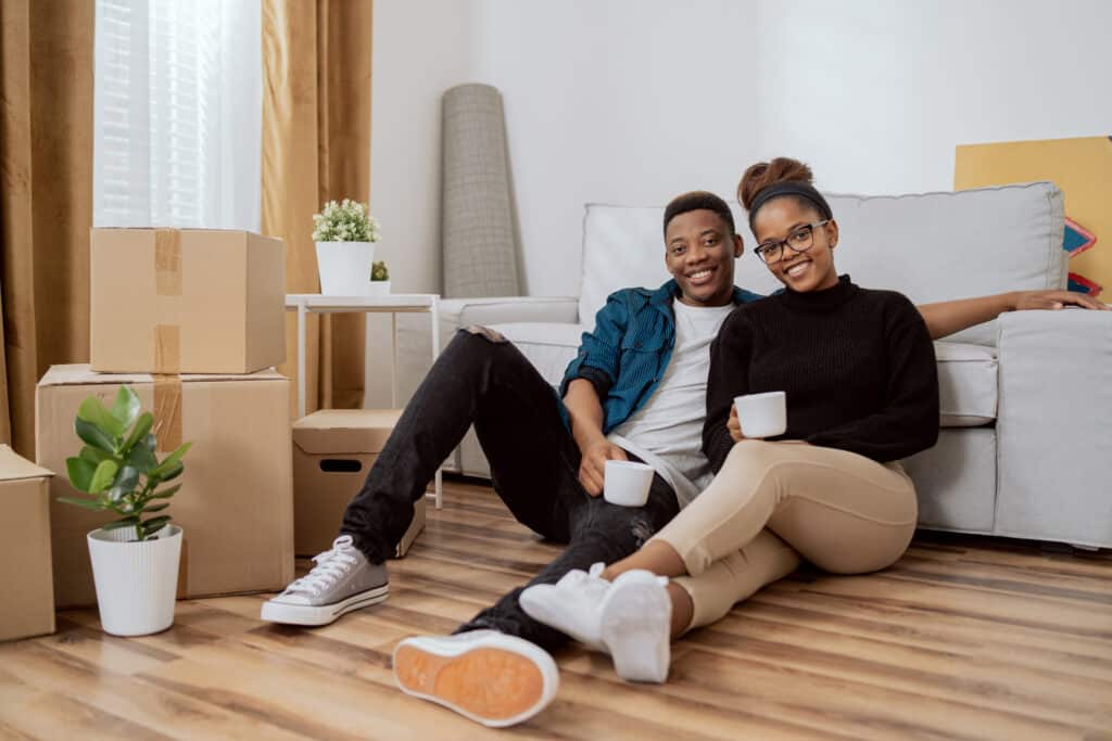 renters insurance. Smiling couple sitting on the floor relaxing after moving in, drinking coffee in their new apartment, around them boxes with unpacked things, the husband embraces his wife