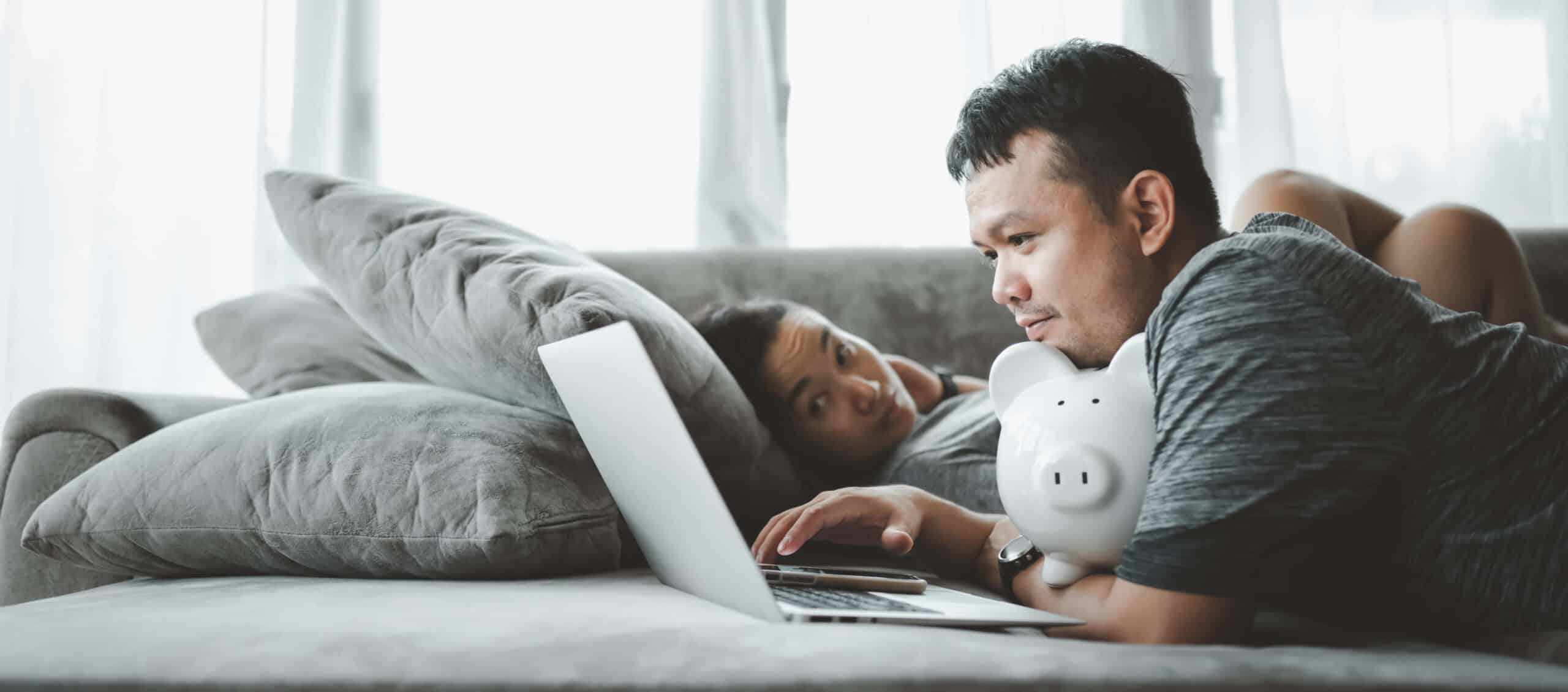 Asian man lying on the couch working on a budget spreadsheet while holding a piggy bank.