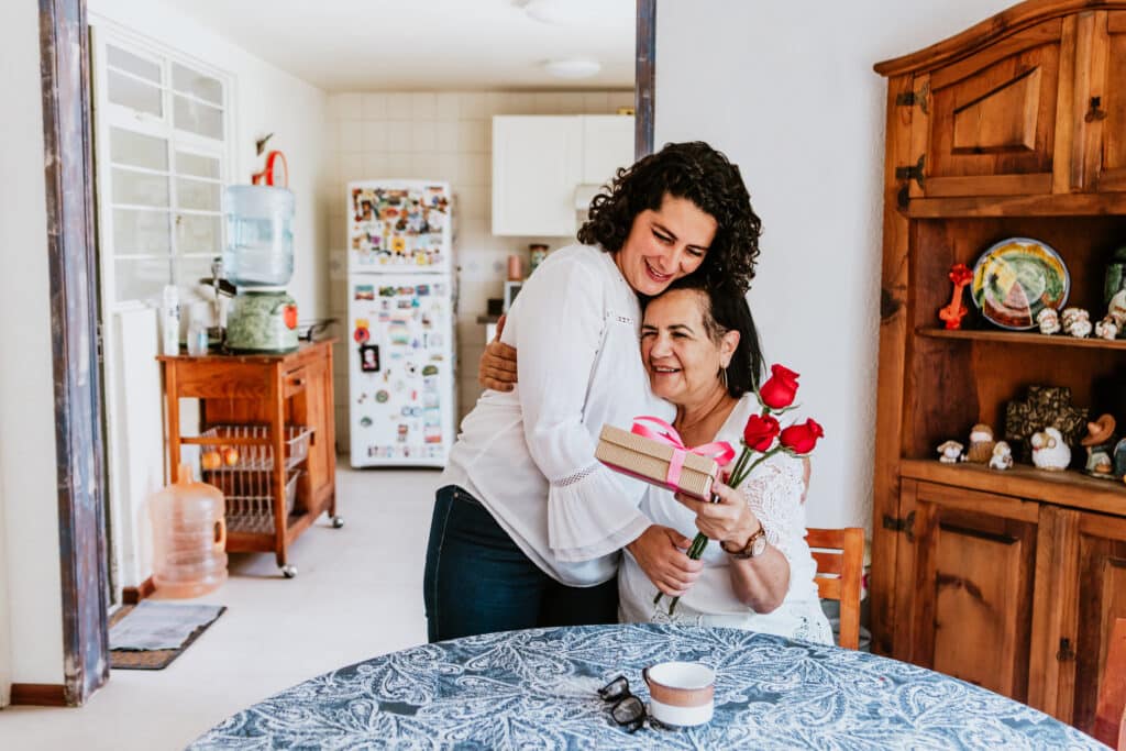 latin young woman and her mother middle age with flowers and gift box at home celebrating Happy mother's day in Mexico city