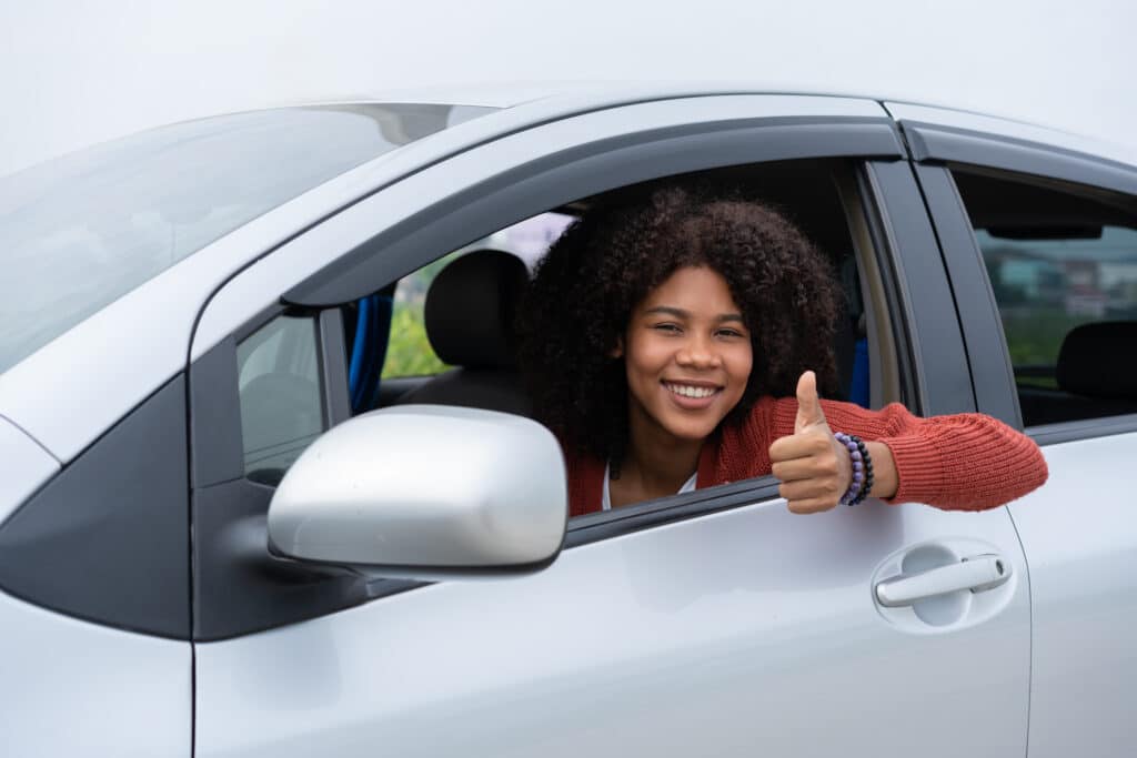 car insurance, young black woman sitting in a silver car giving a thumbs up