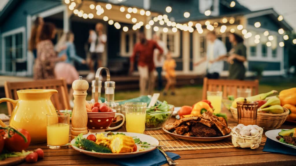 affordable summer BBQ, table with food, lights strung overhead, in a backyard.