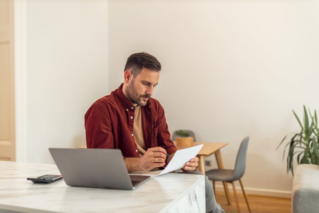 savings account, middle-aged white man with brown hair and beard sitting with a computer looking at a piece of paper.