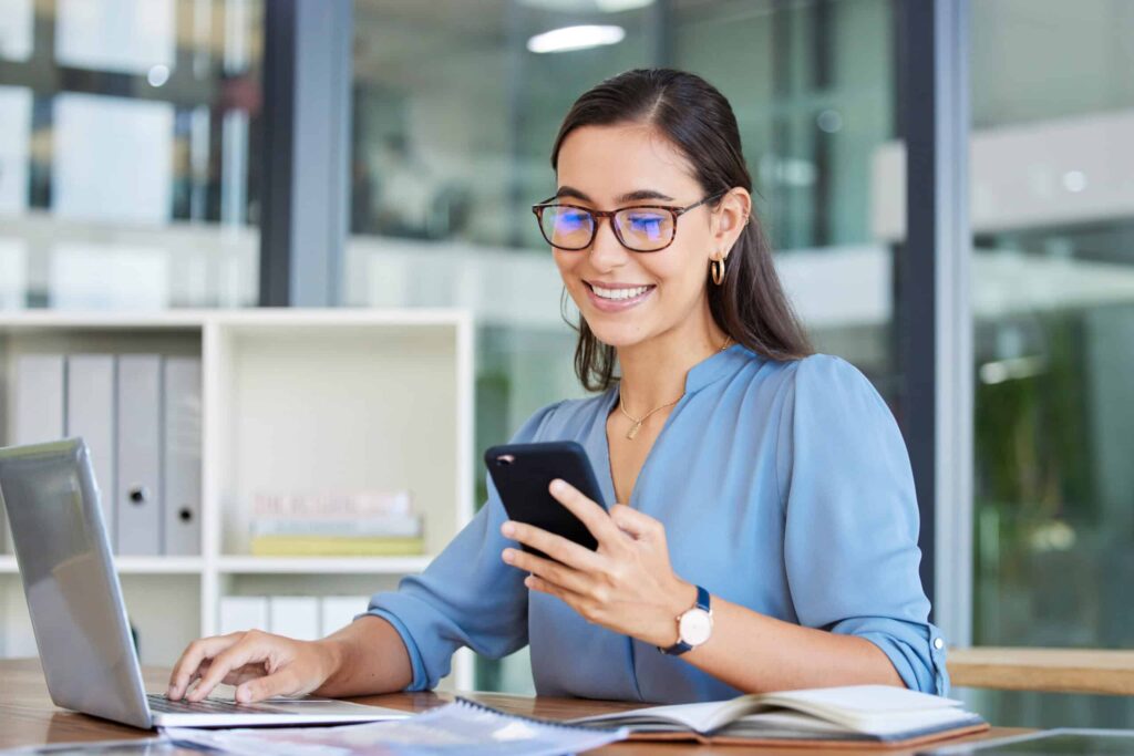 free calling. Business woman, laptop and smile for phone, communication or reading at the office. Happy female employee event planner checking email, social media or post on smartphone and computer at workplace