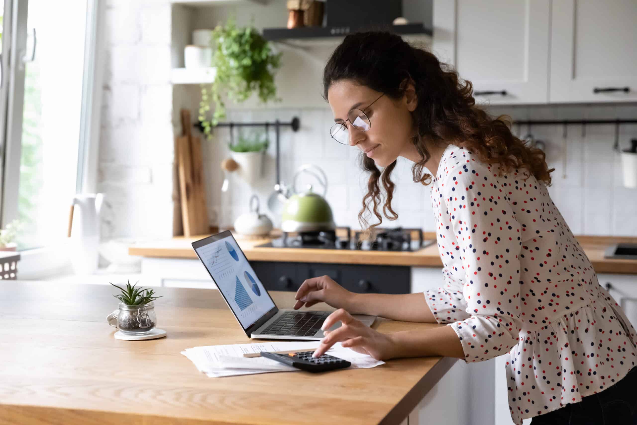 save or invest: Serious young woman wearing glasses calculating finances, household expenses, confident businesswoman working with project statistics, using laptop and calculator, standing in kitchen at home