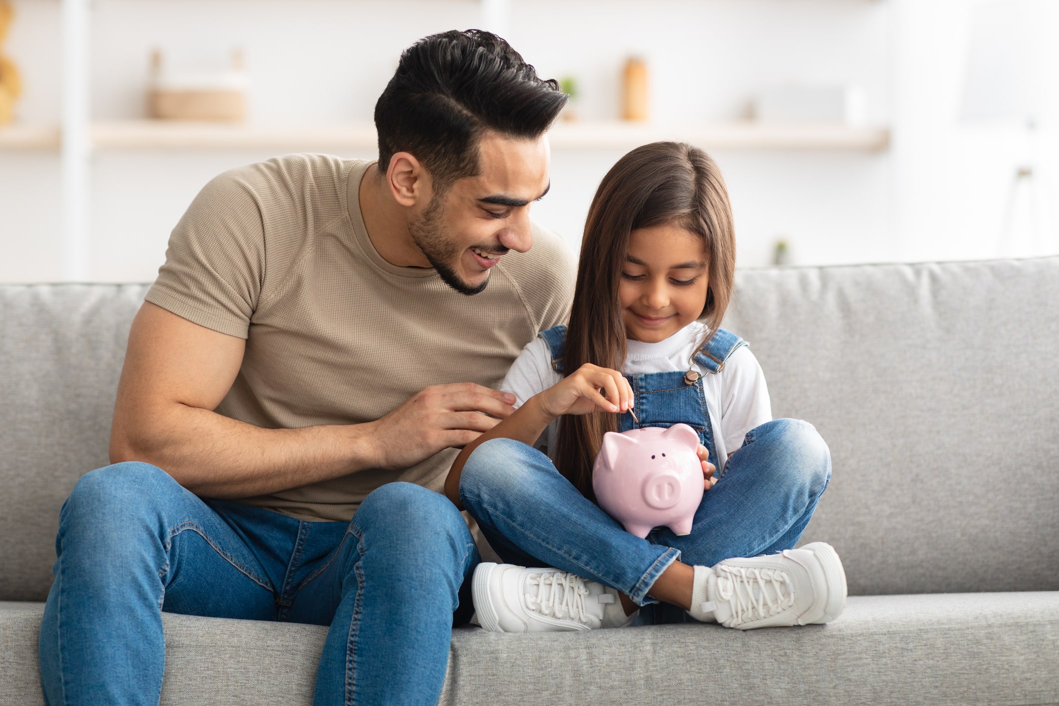 money lessons to teach your kids. Financial Education For Children Concept. Portrait of cute smiling little girl putting coin in pink piggy bank, sitting with dad on the couch at home, man teaching his daughter how to invest.