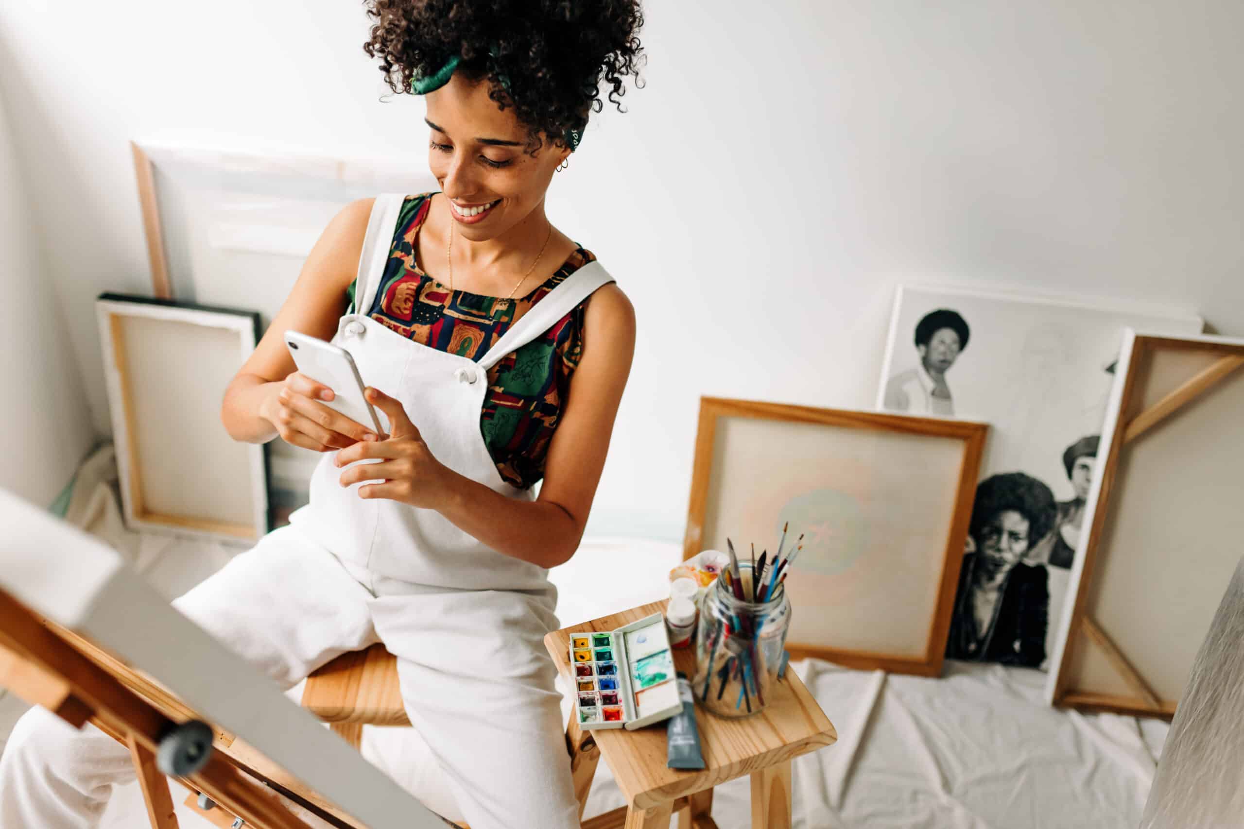 Side hustle taxes: Vlogger taking a picture of her artwork in her workshop. Happy young female painter smiling while capturing her painted canvas with a smartphone. Female freelancer creating an online painting blog.