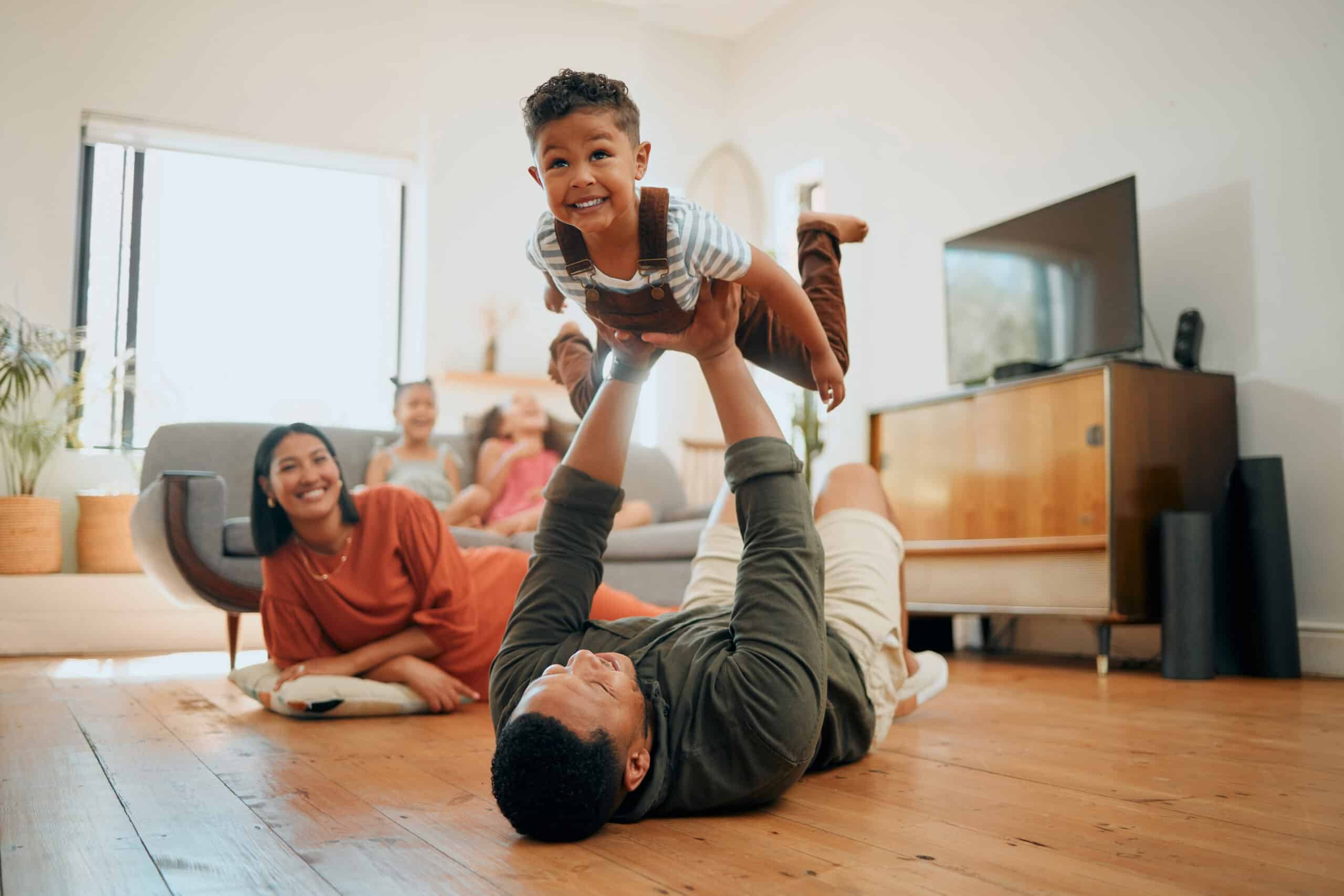 kid costs: A happy mixed race family of five relaxing in the lounge and being playful together. Loving black family bonding with their son while playing fun games on the floor at home