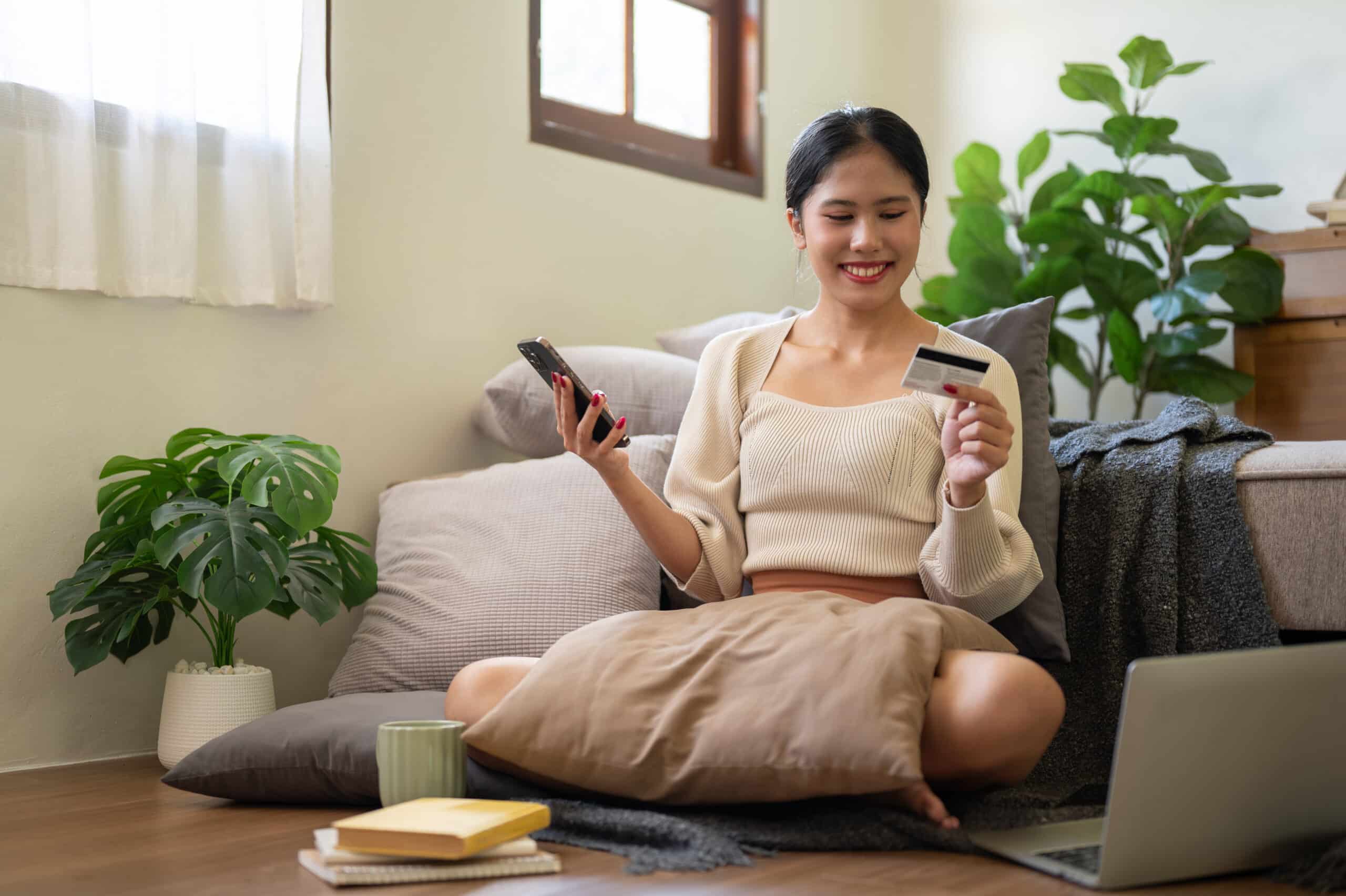 financial minimalism. Happy Asian woman is sitting on the floor beside her bed and enjoying shopping online via her phone.