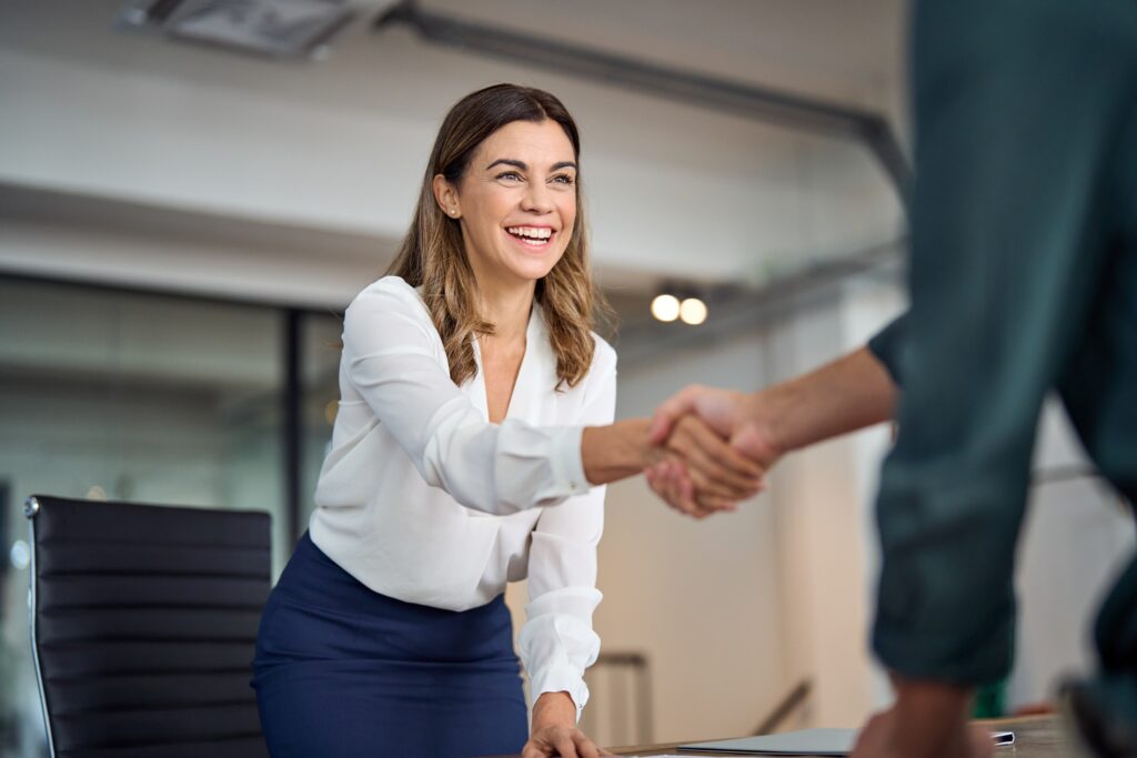 Negotiate. Happy mid aged business woman manager handshaking greeting client in office. Smiling female executive making successful deal with partner shaking hand at work standing at meeting table.