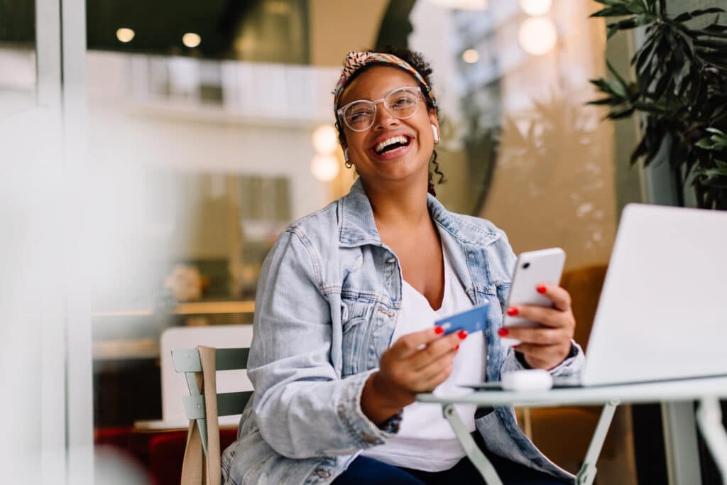 buy happiness. Young woman in a café happily uses her smartphone for online shopping and mobile banking, paying with a credit card. Woman highlighting the convenience of wireless technology and internet banking for digital transactions.