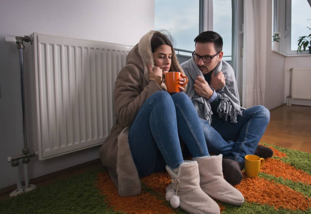 keep warm. Young couple in jacket and covered with blanket sitting on floor beside radiator and trying to warm up