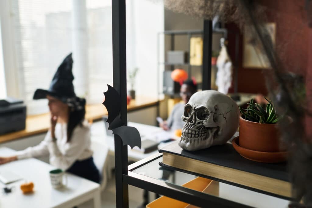 Halloween. Human skull and flowerpot with green domestic plant standing on book on shelf against young businesswoman wearing witch hat