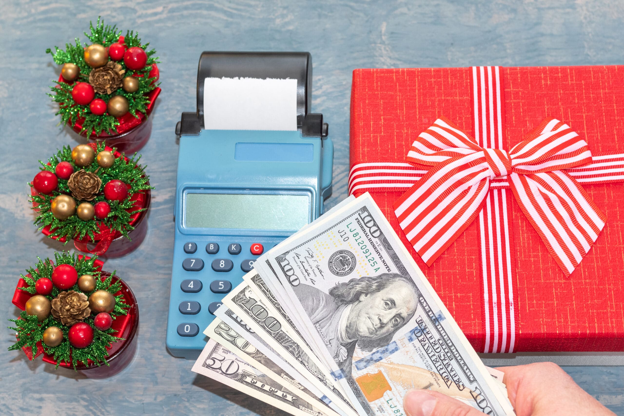 holiday budget. Christmas sale. A hand holding dollars over a cash register near a red gift box with a ribbon and small Christmas trees with cones. online shopping. Buying gifts for New Year and Christmas