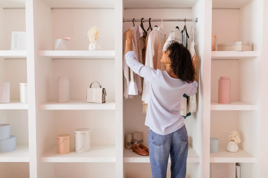 spending. Curly-haired woman in casual attire thoughtfully arranging her stylish clothing in an open white closet in a room with soft pink walls